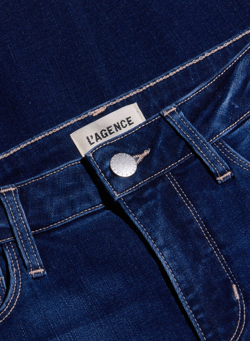 Close-up of dark blue denim jeans showing the waistband, button, belt loops, and a white fabric label that reads 'L'AGENCE'.