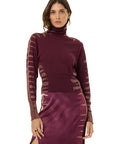 A woman modeling a long-sleeve turtleneck dress in deep plum purple, featuring sheer patterned sleeves and a satin-like skirt, against a plain white background.