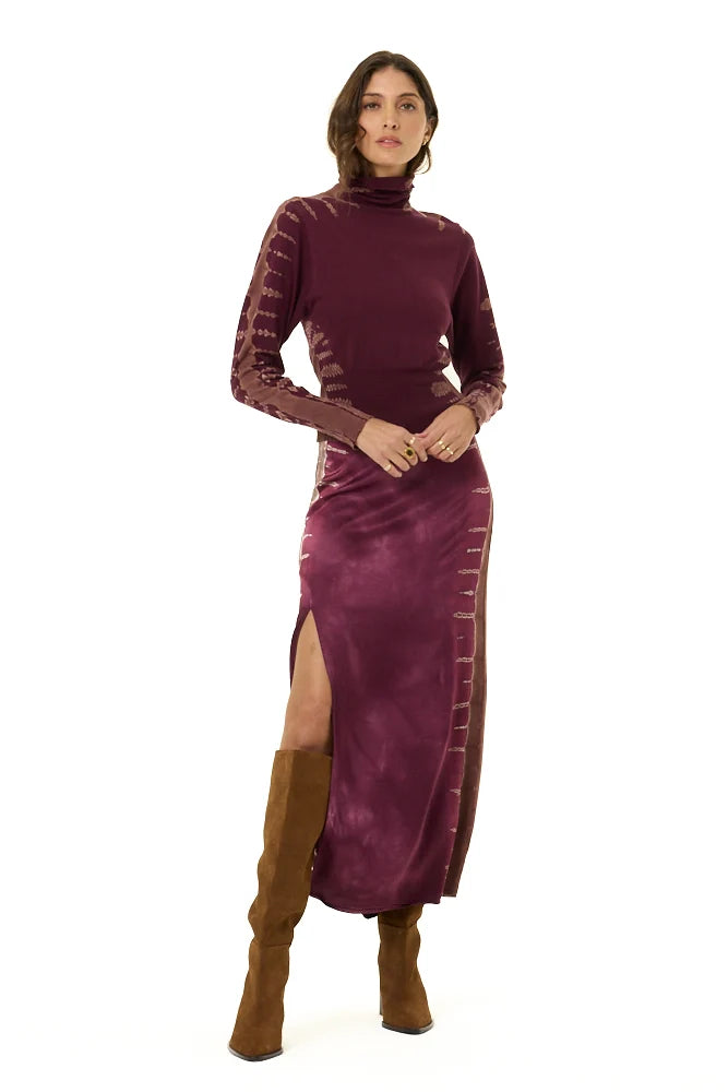 A model wearing a long-sleeved, high-neck plum velvet midi dress with side slits and brown suede knee-high boots, photographed against a solid white background.