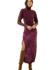 A model wearing a long-sleeved, high-neck plum velvet midi dress with side slits and brown suede knee-high boots, photographed against a solid white background.