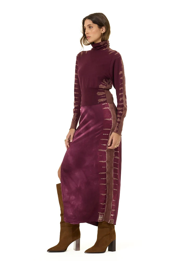 A woman modeling a long-sleeve, high-neck maxi dress in a deep plum purple with textured vertical stripes down the sleeves and sides, paired with brown knee-high boots, against a plain white background.