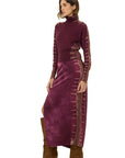 A woman modeling a long-sleeve, high-neck maxi dress in a deep plum purple with textured vertical stripes down the sleeves and sides, paired with brown knee-high boots, against a plain white background.