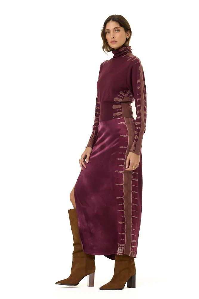 A model wearing a long-sleeved, high-neck maxi dress in deep plum purple velvet with metallic gold embroidered details down the sleeves and sides, paired with brown suede knee-high boots, against a plain white background.