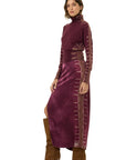 A model wearing a long-sleeved, high-neck maxi dress in deep plum purple velvet with metallic gold embroidered details down the sleeves and sides, paired with brown suede knee-high boots, against a plain white background.
