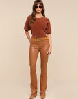 A woman modeling a brown short-sleeve knit top and brown coated jeans with a tan belt, photographed against a plain beige studio background.