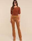 A woman modeling a brown short-sleeve knit top and matching brown pants with a wide brown belt, photographed against a plain beige studio background.