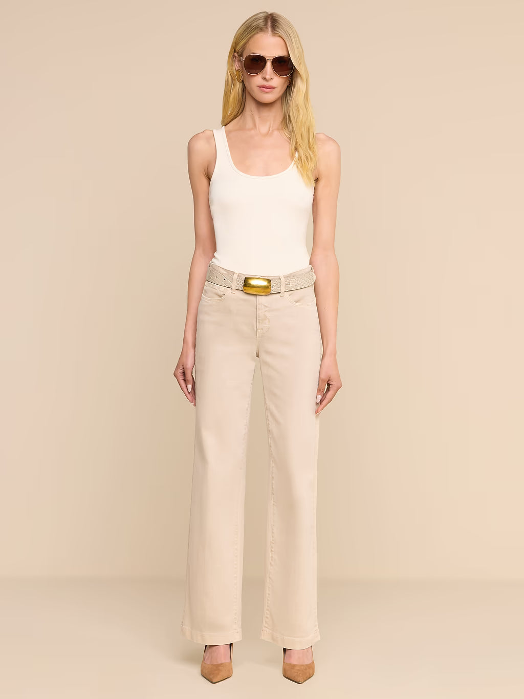 A blonde model wearing a white ribbed tank top, beige wide-leg jeans with a beige belt featuring a gold buckle, and beige heels, standing against a solid beige studio background.