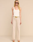 A blonde model wearing a white ribbed tank top, beige wide-leg jeans with a beige belt featuring a gold buckle, and beige heels, standing against a solid beige studio background.