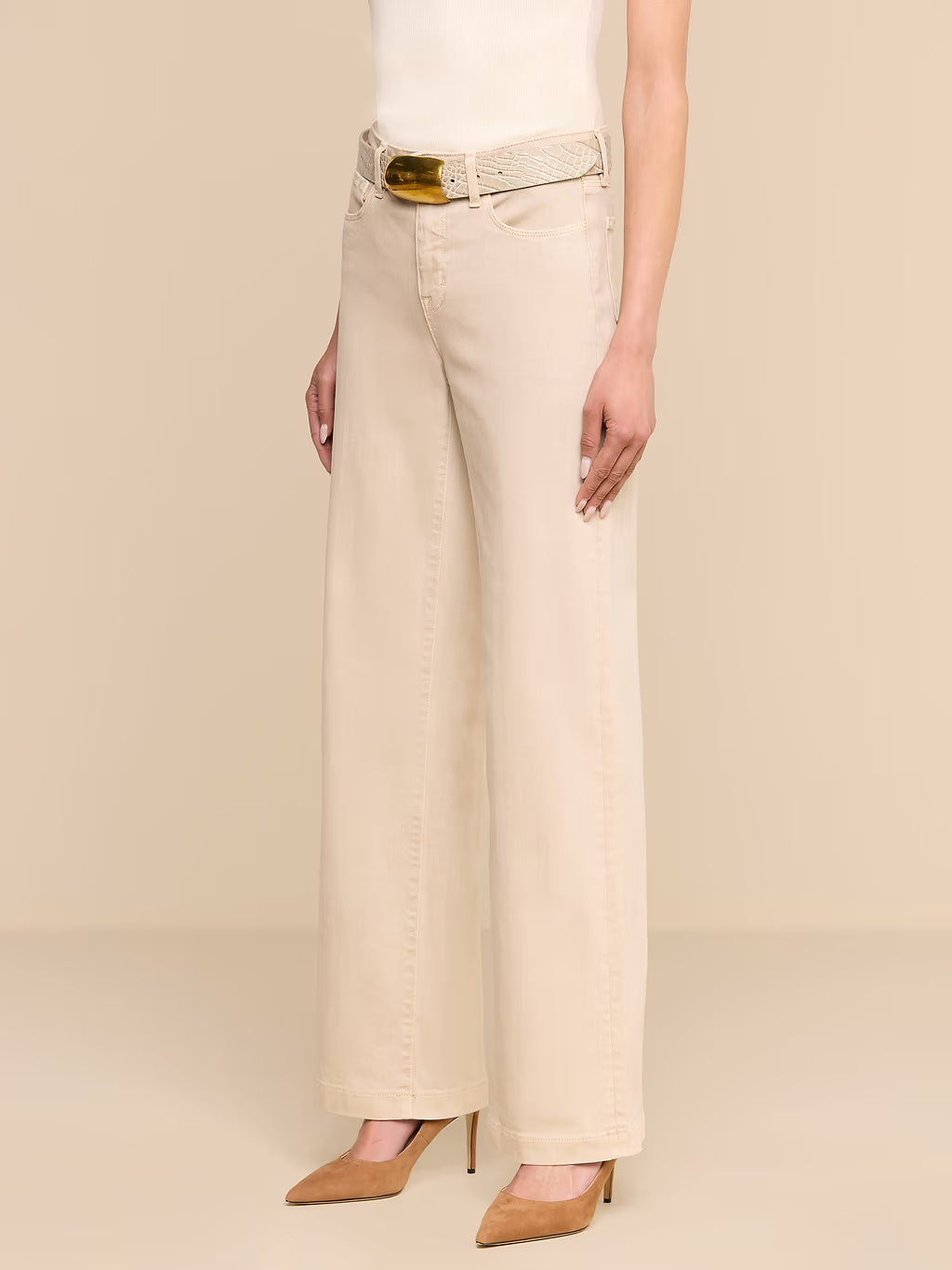 A model wearing a pair of light beige wide-leg jeans with a matching belt featuring a gold buckle, paired with beige pointed-toe heels against a solid beige background.
