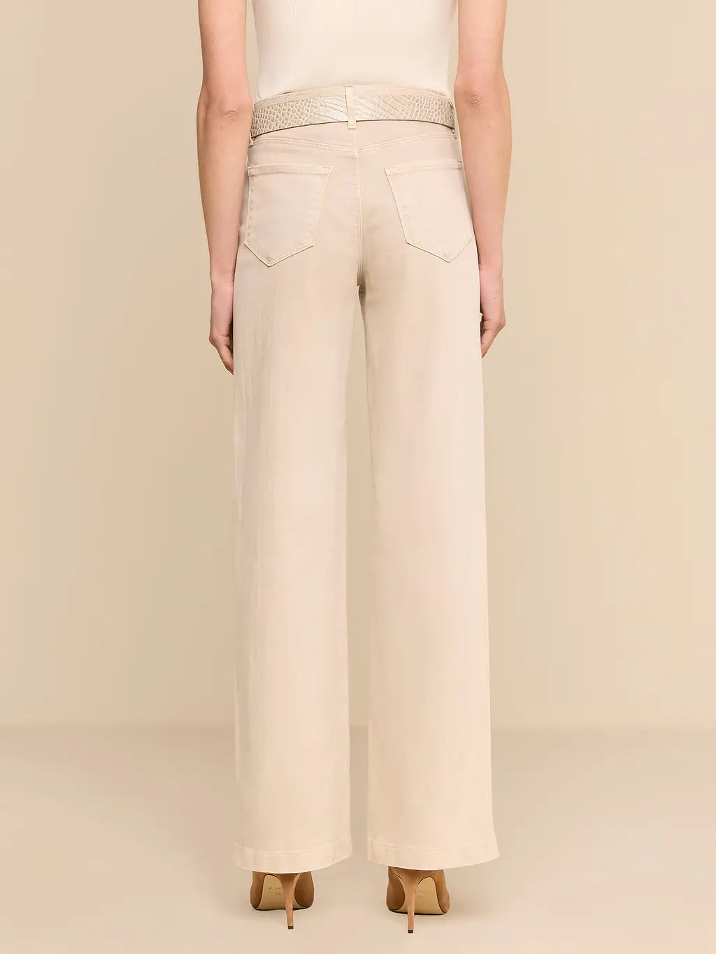 A model shown from the back wearing high-waisted, wide-leg trousers in a light cream or beige color, paired with a textured belt and pointed-toe heels against a solid beige studio background.