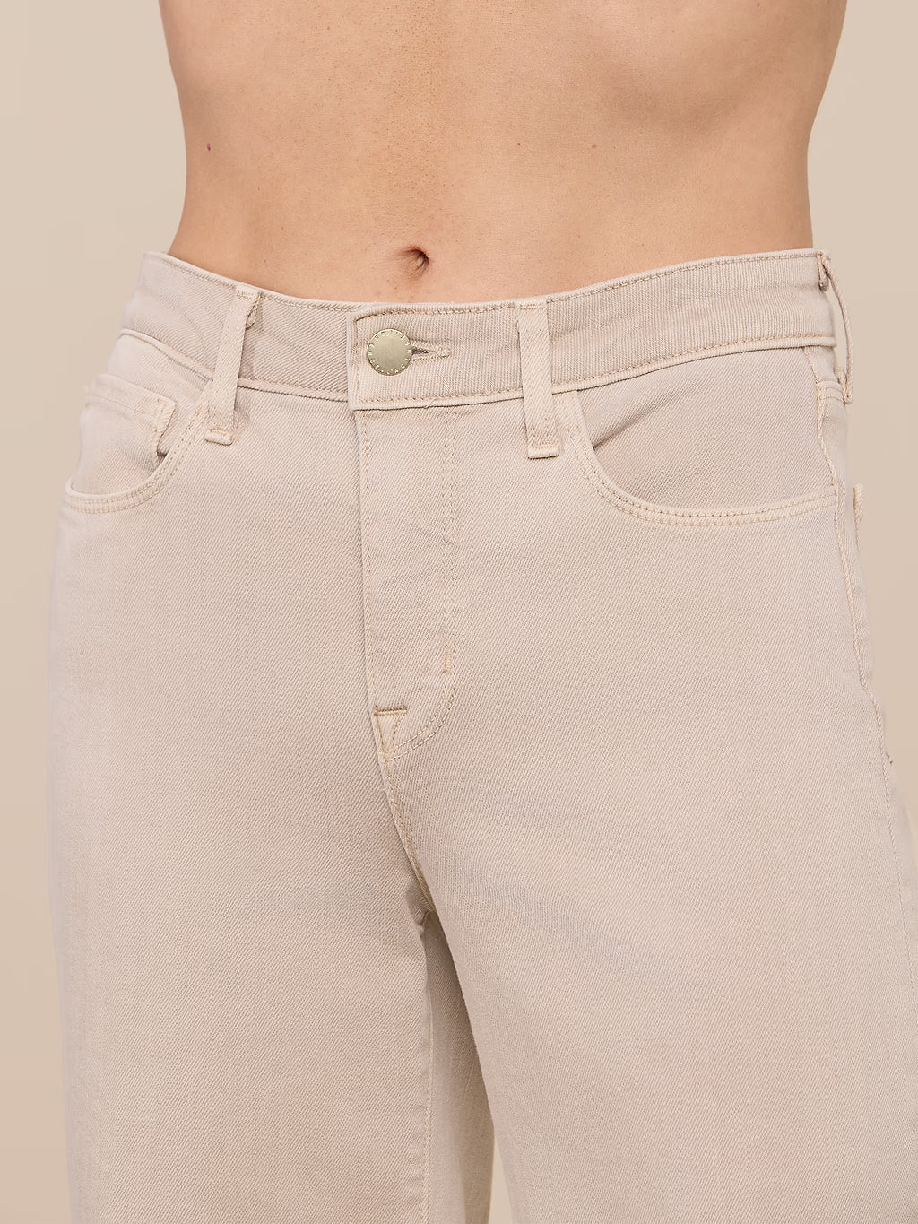 Close-up of a model's lower torso wearing light beige denim jeans against a solid beige background.