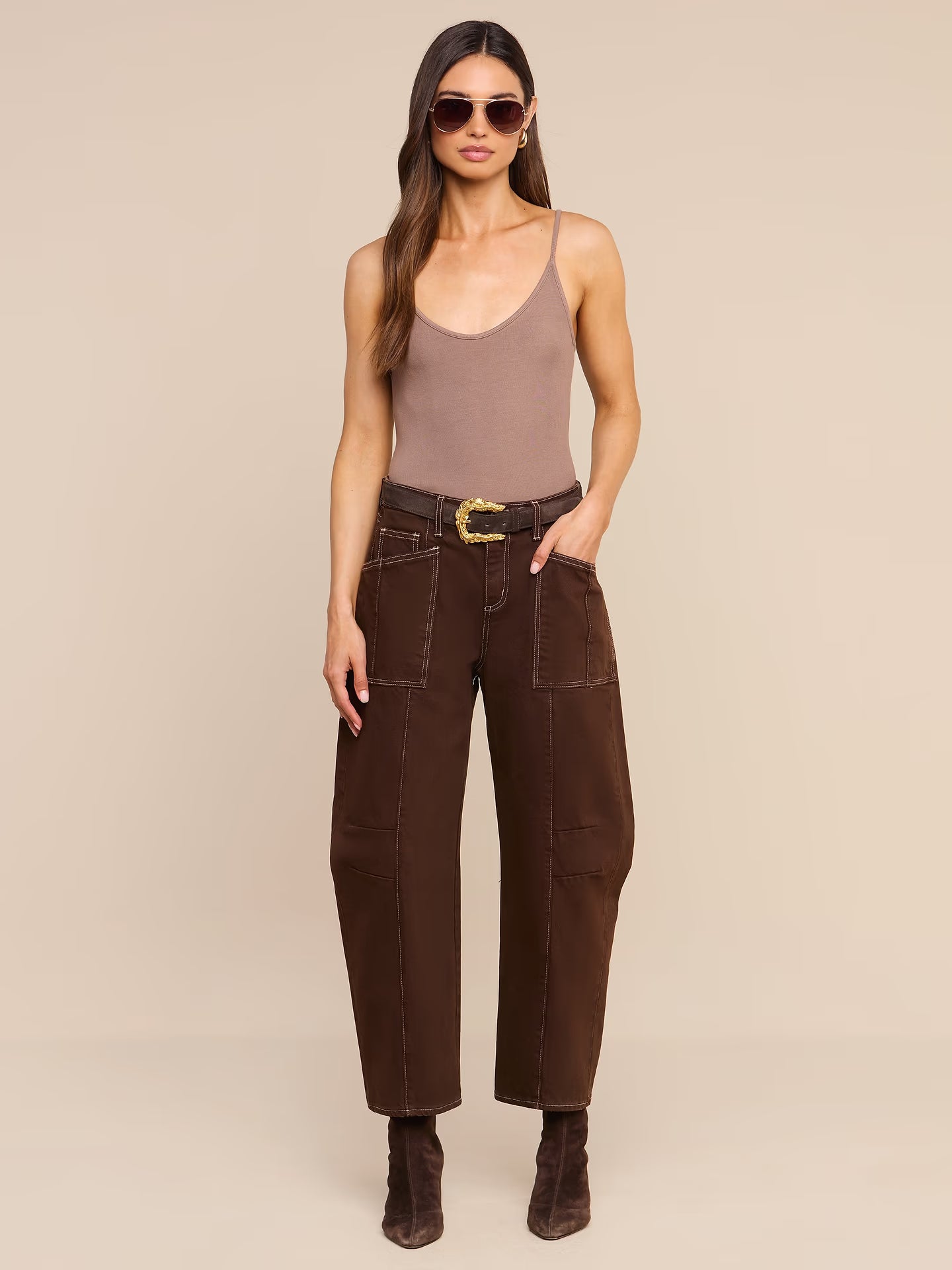 A woman modeling a pair of dark brown wide-leg cropped pants with a matching brown belt featuring a gold buckle, styled with a taupe camisole, sunglasses, and brown ankle boots against a solid beige background.
