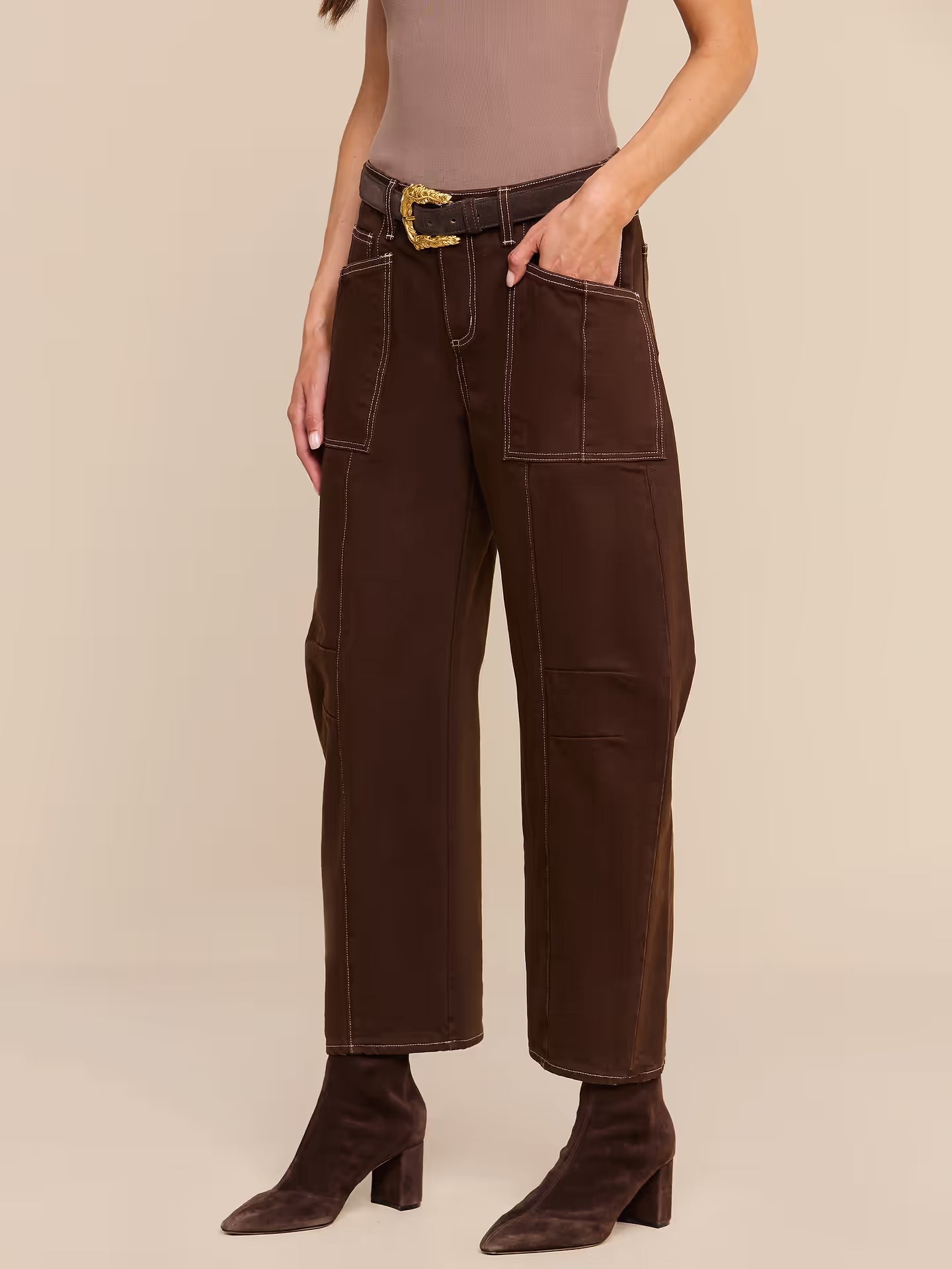 A model wearing dark brown wide-leg cropped pants with a matching dark brown belt featuring a gold buckle, styled with a mauve top and dark brown heeled ankle boots against a solid beige studio background.
