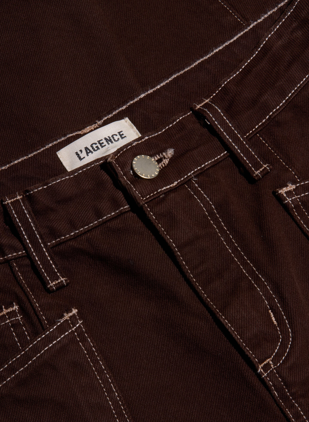Close-up of dark brown corduroy pants showing the waistband, belt loops, button, and white contrast stitching. A white fabric label reading 'L'AGENCE' is visible near the waistband.