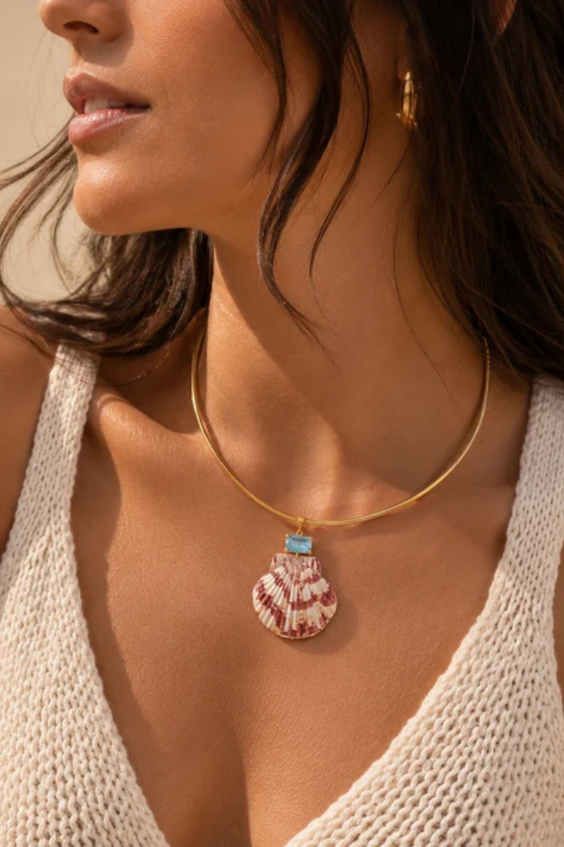 A close-up lifestyle shot of a woman wearing a delicate gold bangle necklace with a seashell-shaped pendant and a small turquoise accent, paired with a white knit top.