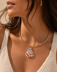 A close-up lifestyle shot of a woman wearing a delicate gold bangle necklace with a seashell-shaped pendant and a small turquoise accent, paired with a white knit top.
