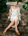 A woman sits on rustic stone steps outdoors, wearing a cream satin slip dress with lace trim, a matching cream knit cardigan, and tan suede flats. Lush green foliage frames the stone pathway.