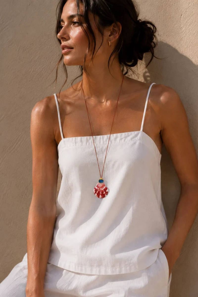 A woman wearing a white spaghetti-strap camisole top and matching bottoms, styled with a long red and blue \pendant necklace, leaning against a textured beige wall.