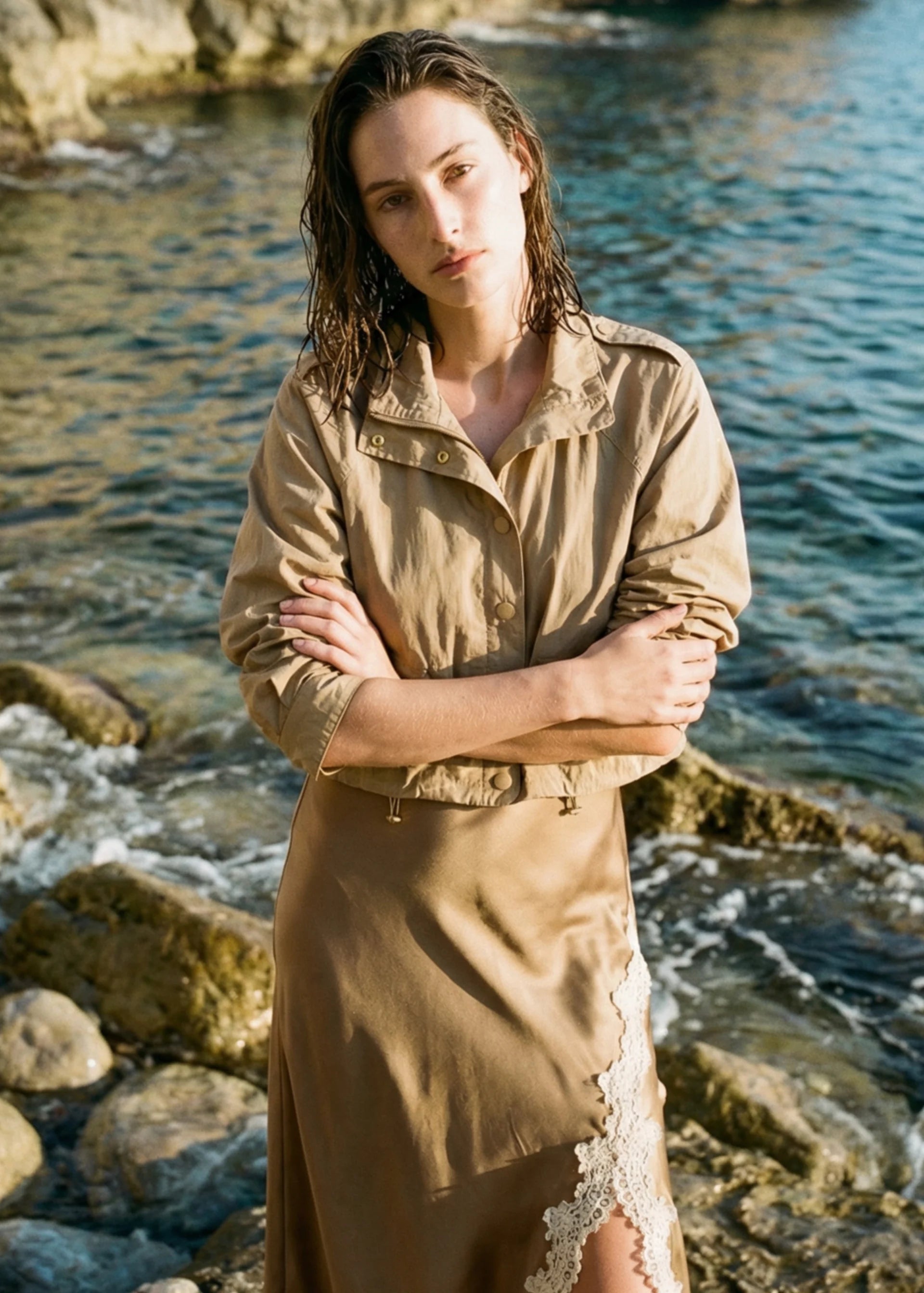 A woman stands on a rocky shoreline with water behind her, wearing Kellen Jacket