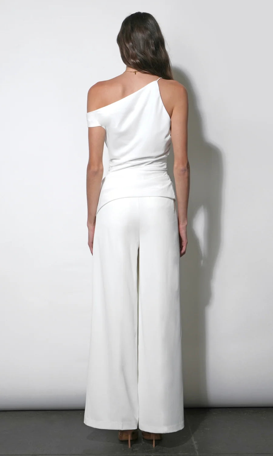 A woman shown from the back wearing a white one-shoulder wrap-style jumpsuit with wide-leg pants, standing against a plain light gray wall.