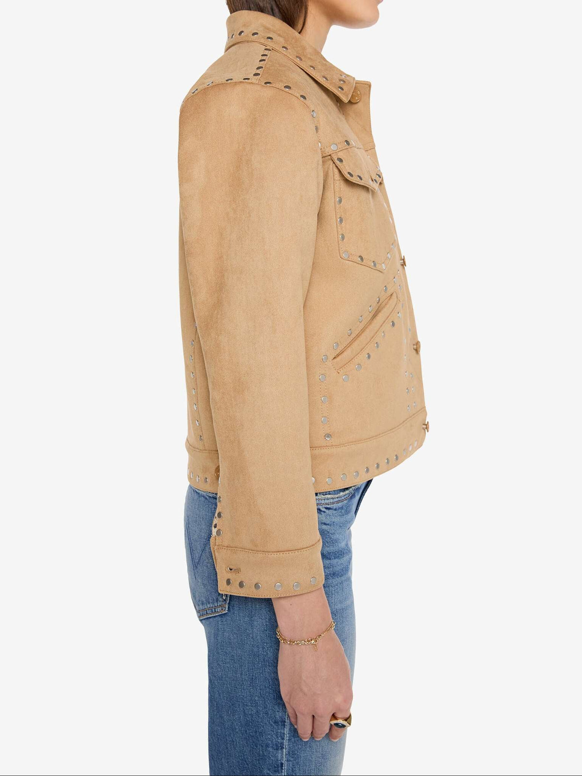 A woman shown in side profile wearing a cropped tan suede jacket with silver studs and blue jeans, photographed against a plain light gray studio background.