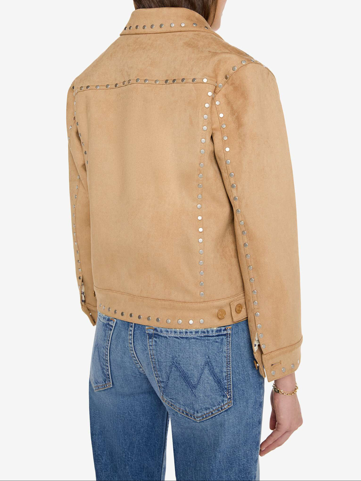 A person shown from the back wearing a tan suede jacket with silver stud detailing and blue jeans, photographed against a plain light gray studio background.
