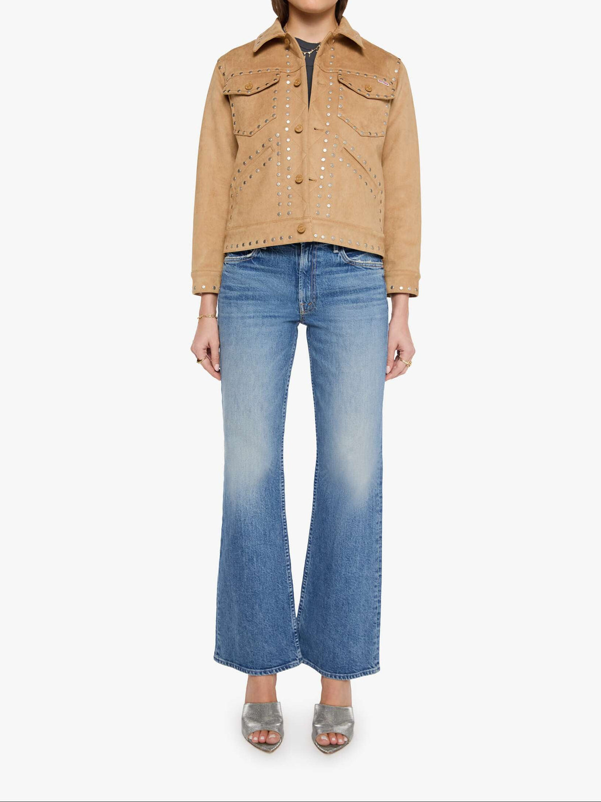 A model wearing a tan suede studded jacket, blue flare jeans, and silver heeled sandals, standing against a plain white studio background.