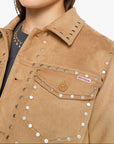 A woman wearing a tan suede jacket with silver studs and shield-style sunglasses, photographed against a clean light gray studio background.