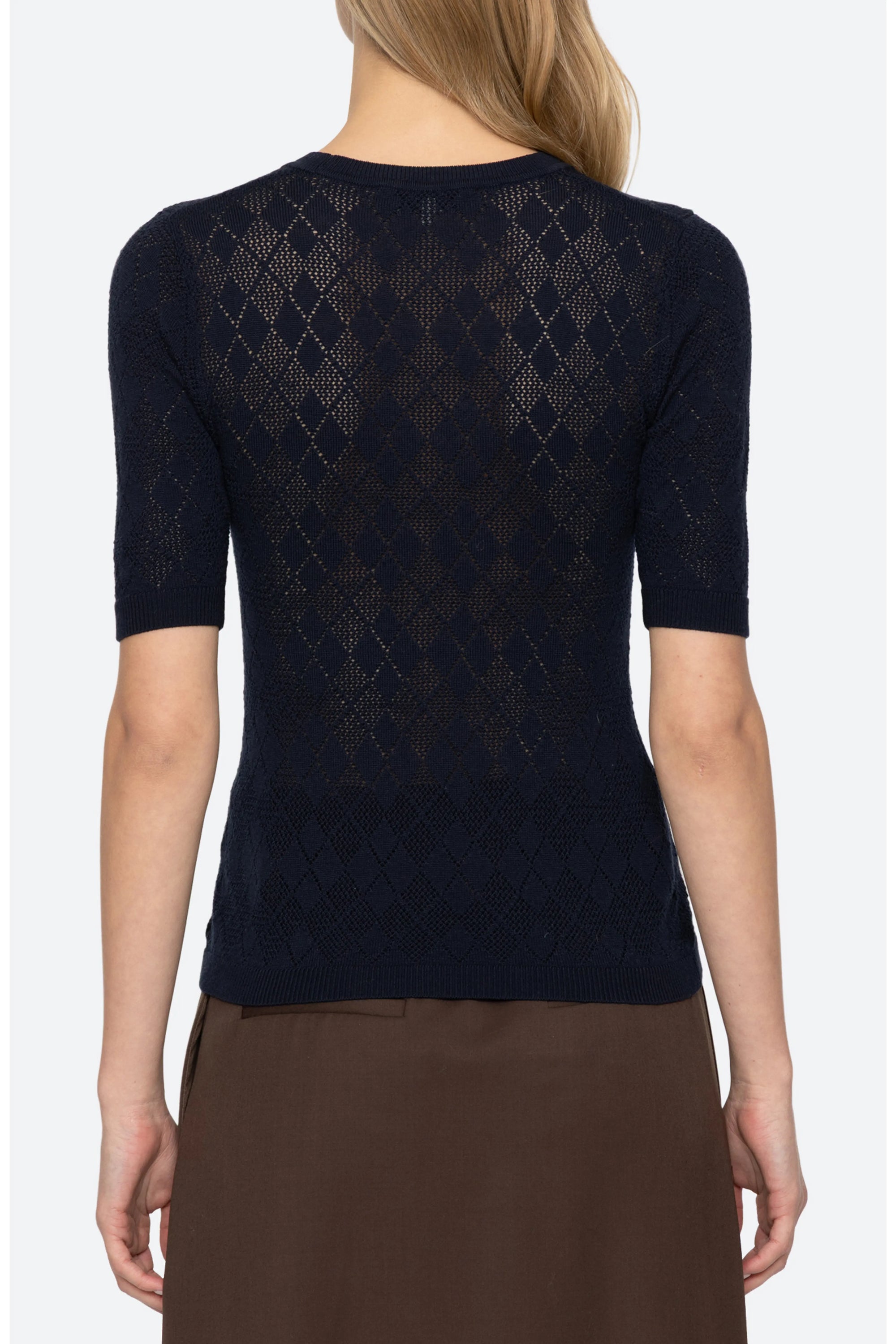 A woman shown from the back wearing a dark navy short-sleeve knit top with an openwork diamond pattern and brown bottoms, against a light gray studio background.