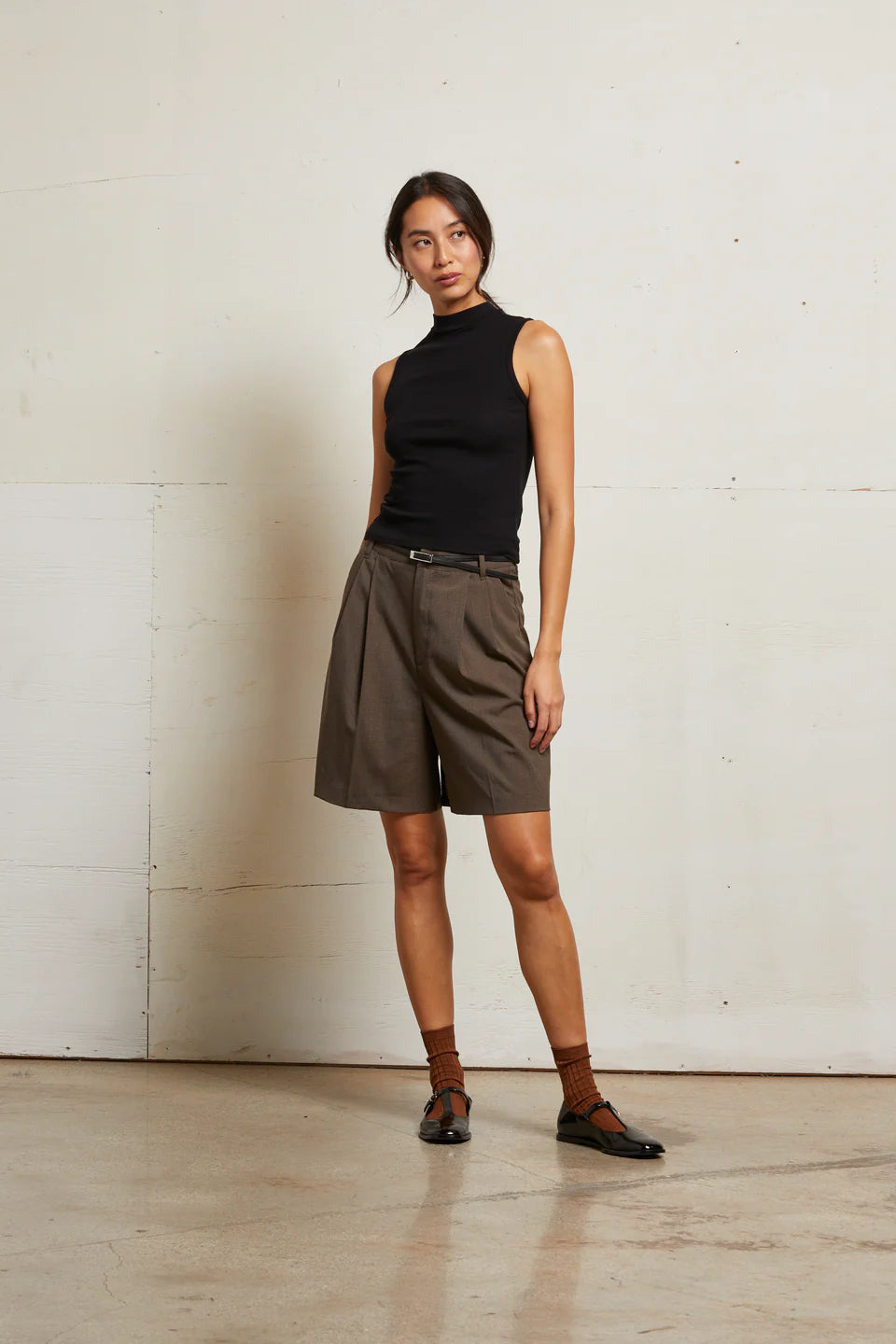 Woman wearing a black sleeveless top and brown shorts against a white wall front view