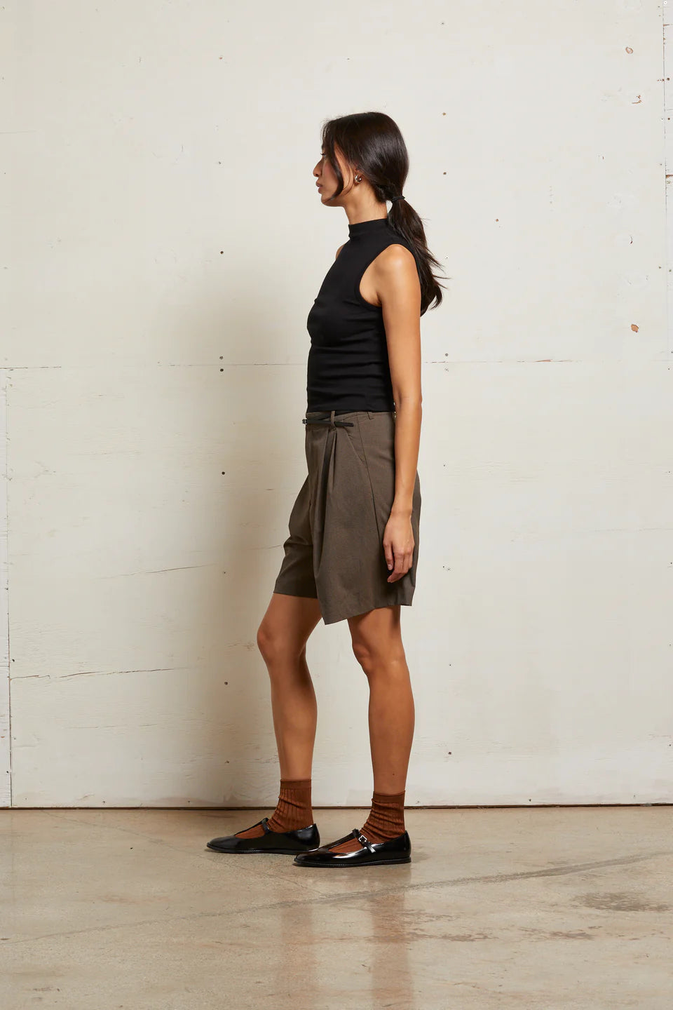 Woman wearing a black sleeveless top and brown shorts against a white wall side view