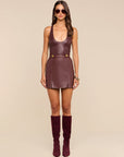 A model wearing Nolina Mini Skirt a dark burgundy faux leather and Tullia Scoopneck Tank, styled with matching burgundy knee-high boots and gold-tone belt hardware, set against a solid beige studio background.