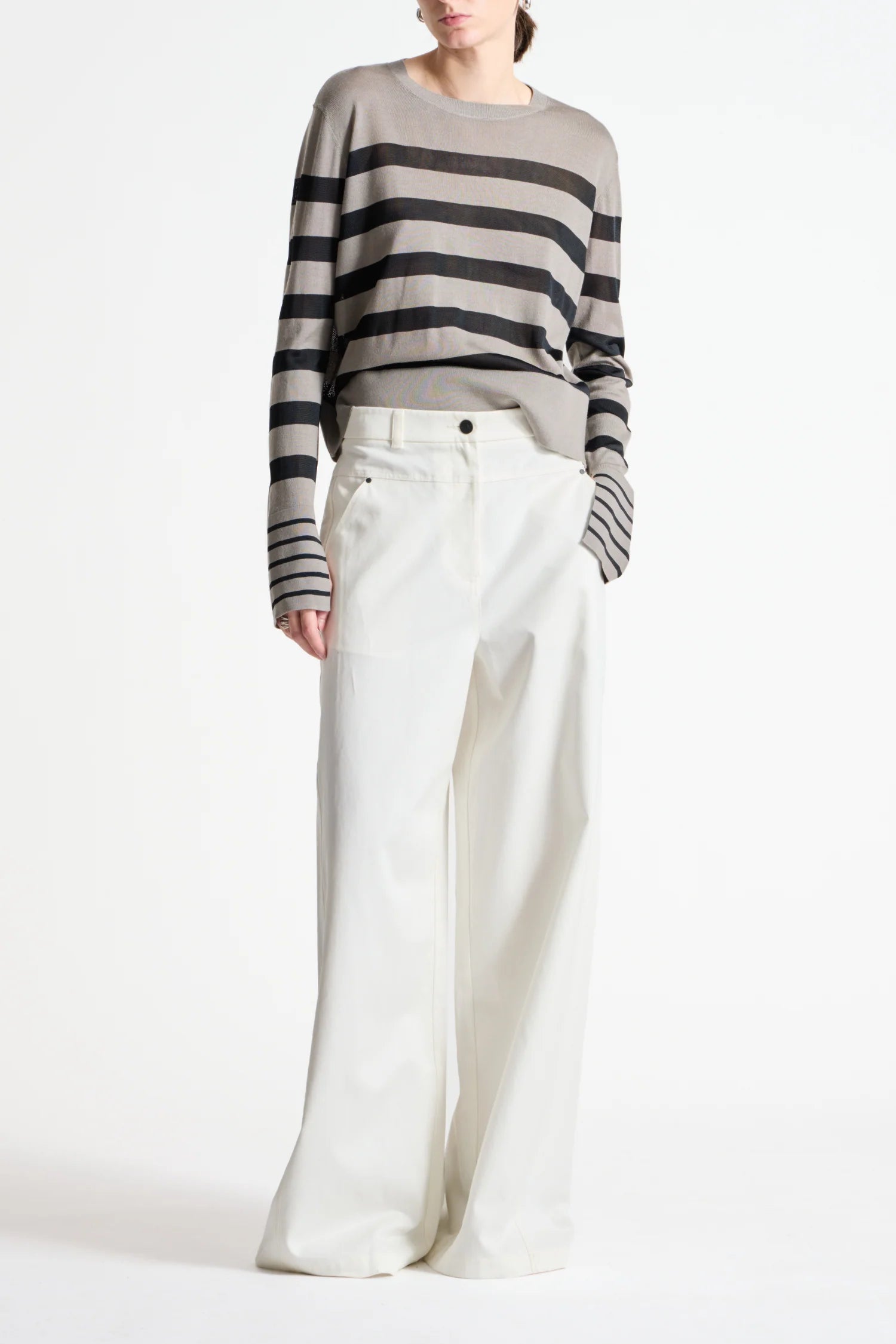 A model wearing a gray and black horizontally striped long-sleeve knit sweater paired with high-waisted, wide-leg white trousers, shot against a plain white studio background.