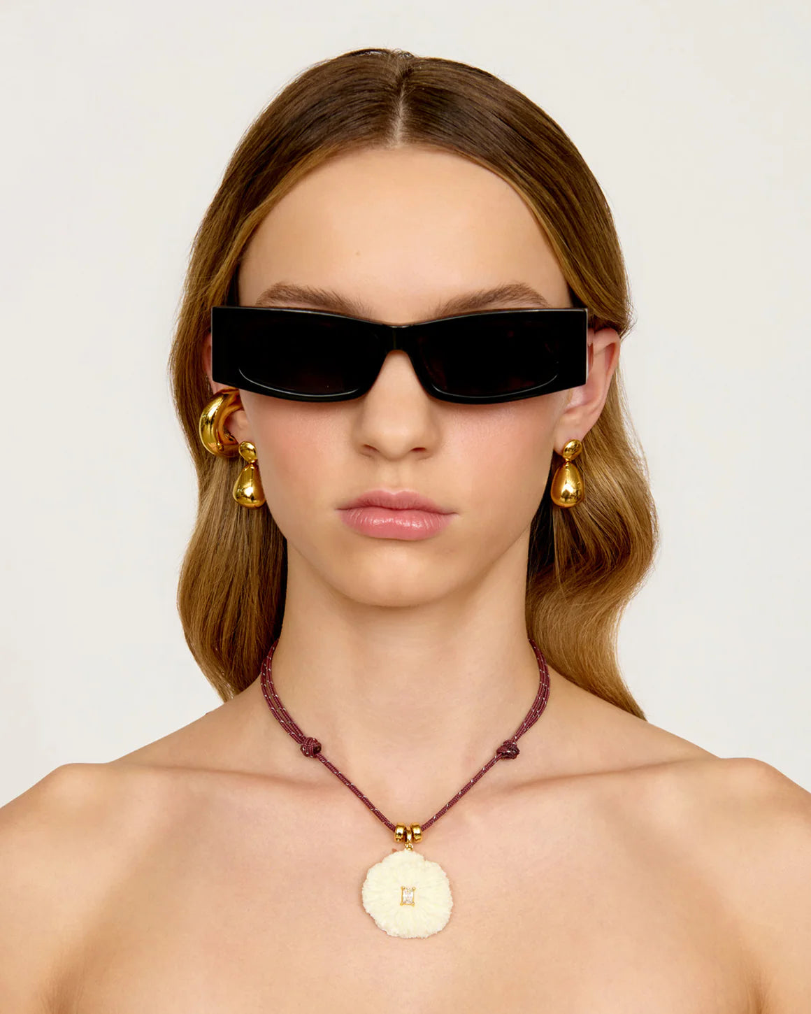 Woman wearing black sunglasses, gold earrings, and a necklace with a shell pendant on a plain background