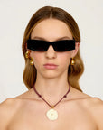 Woman wearing black sunglasses, gold earrings, and a necklace with a shell pendant on a plain background