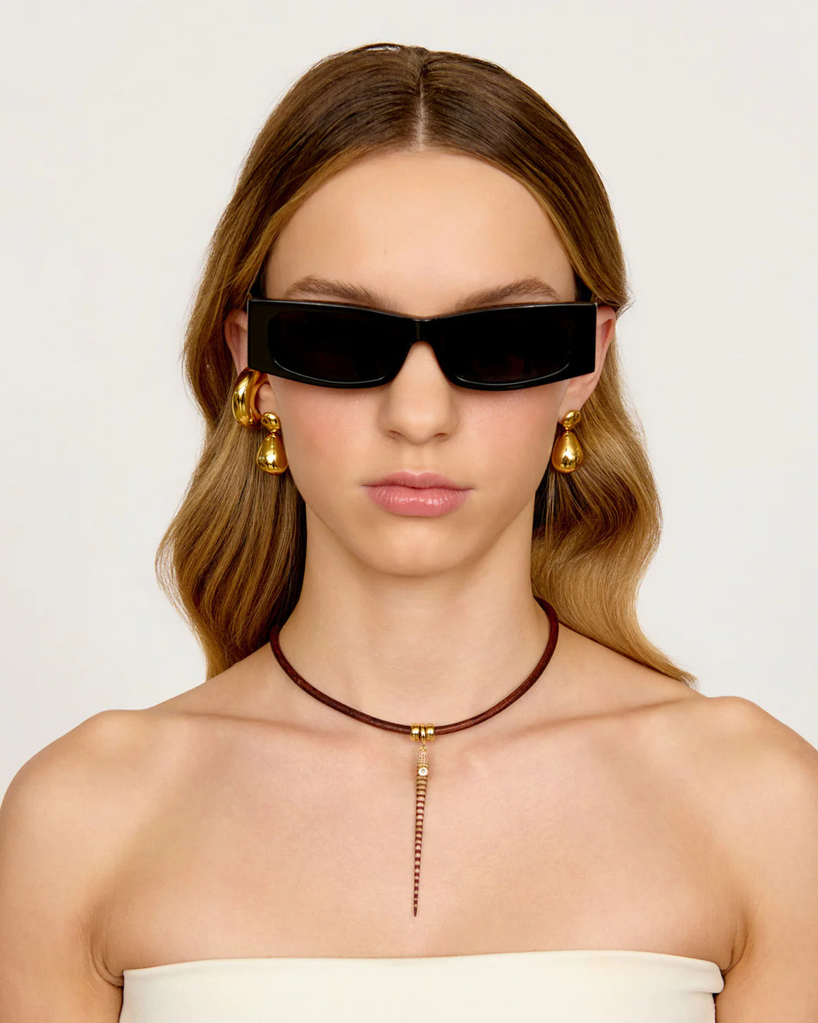 Woman wearing black sunglasses, gold earrings, and a brown leather necklace on a white background