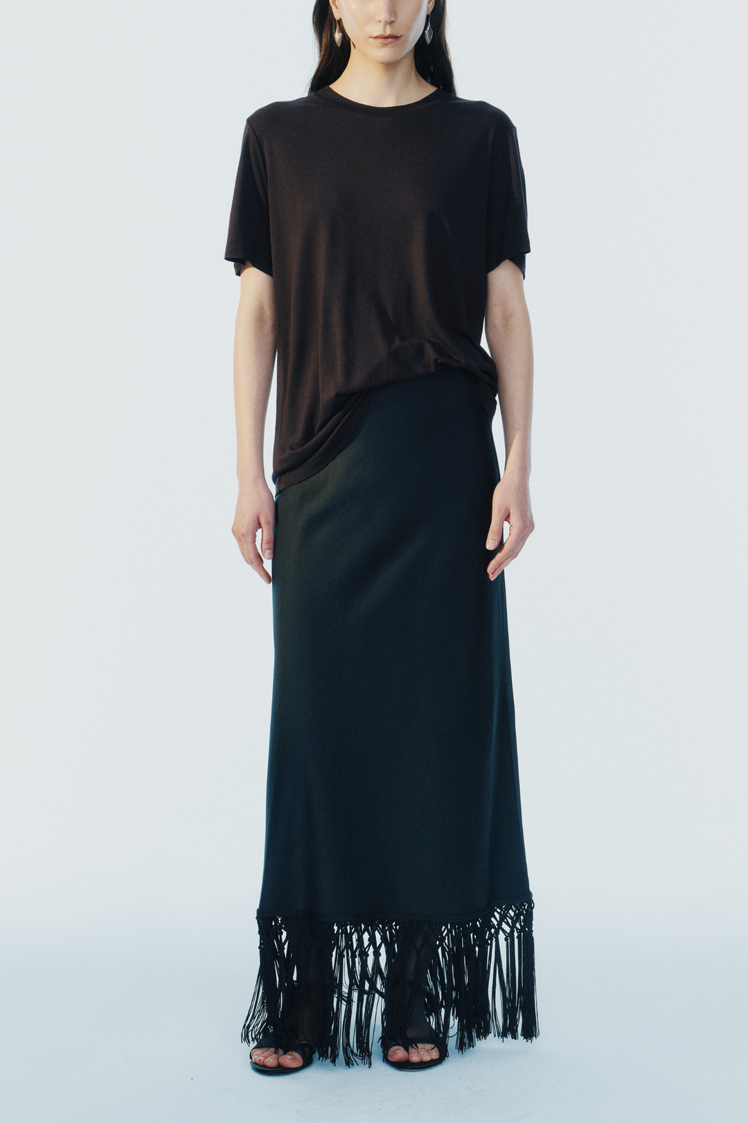 model wearing a black maxi skirt with fringe trim and a short sleeve brown tee on a white background