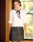 A woman wearing a white blouse with a black bow tie collar and a black faux-leather mini skirt, standing in front of a window with warm light.