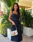 A woman wearing a dark denim midi dress with spaghetti straps and a sweetheart neckline, holding a white patterned handbag. She is standing outdoors in front of large green potted plants and a beige building wall.