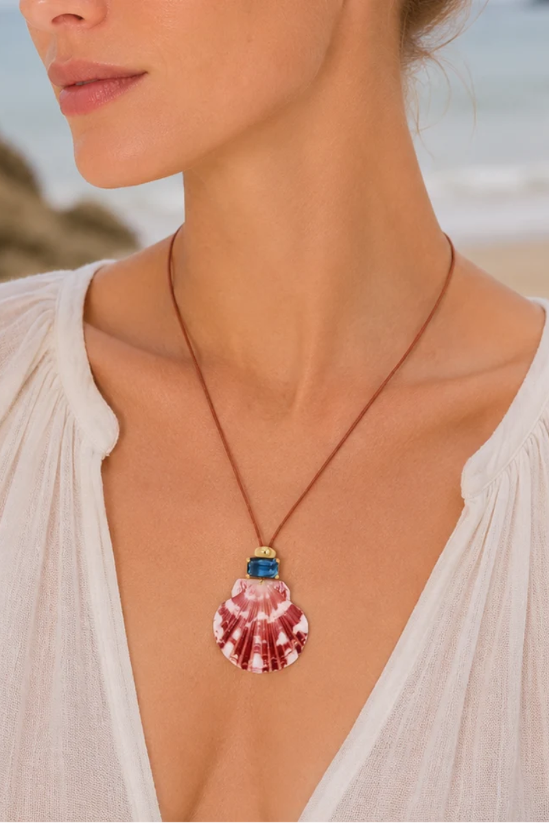 A close-up lifestyle shot of a woman wearing a seashell pendant necklace on a beach. The necklace features a pink and white striped seashell pendant with a gold cap and a small blue stone accent at the top, suspended from a brown cord.