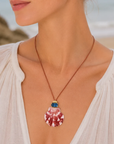 A close-up lifestyle shot of a woman wearing a seashell pendant necklace on a beach. The necklace features a pink and white striped seashell pendant with a gold cap and a small blue stone accent at the top, suspended from a brown cord.