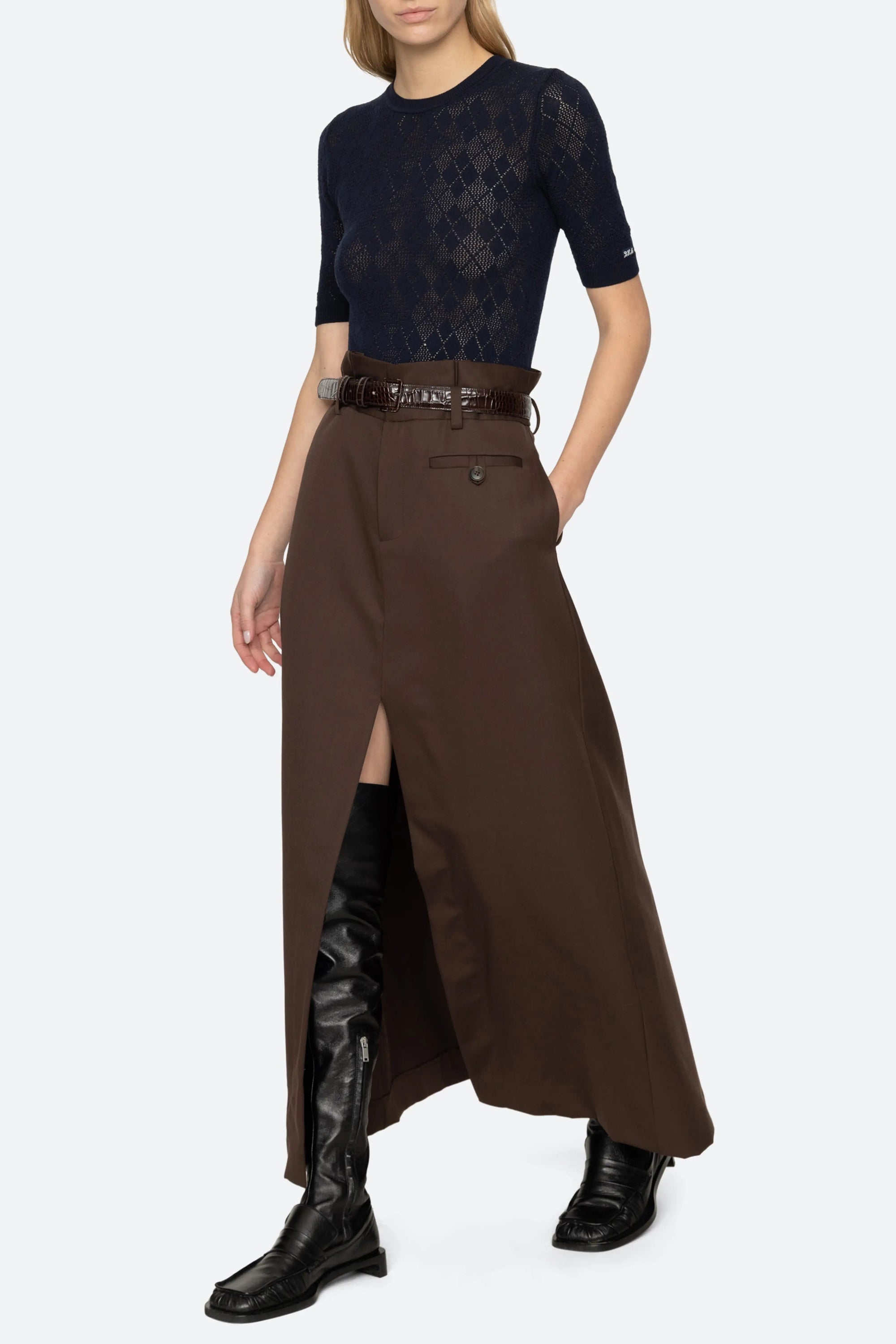 A model stands against a light gray studio background wearing a dark navy short-sleeve lace top, a long brown skirt with a front slit and belt, black knee-high boots, and black ankle boots.