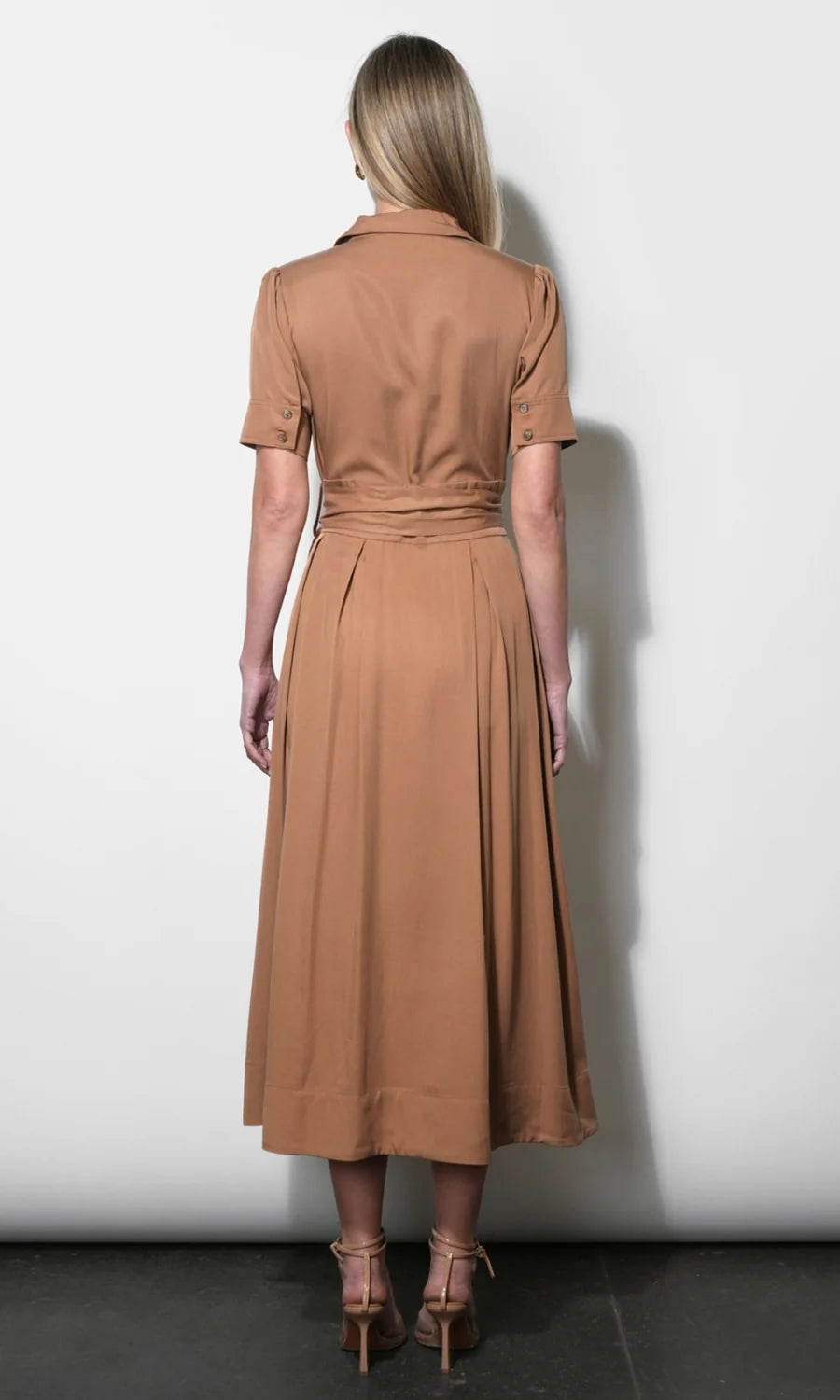 A woman viewed from the back wearing a tan short-sleeve midi dress with a collared neckline and a tied waist, standing against a plain light gray wall.