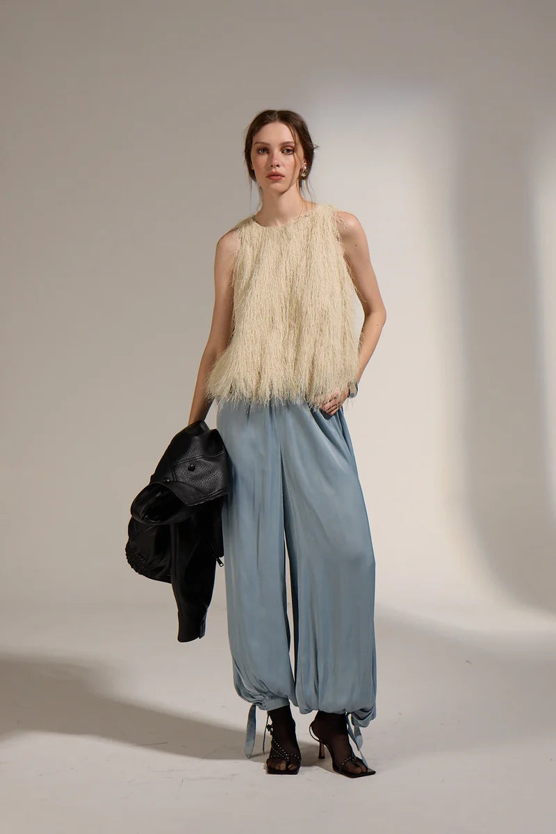 A model stands in a studio wearing a sleeveless cream shaggy top, light blue wide-leg pants, and dark brown heeled boots, while holding a black jacket against a plain light background.