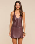 A woman modeling a matching two-piece outfit made of smooth faux leather, featuring a deep V-neck sleeveless top and a high-waisted mini skirt, in a dusty plum-brown color, set against a solid beige background.