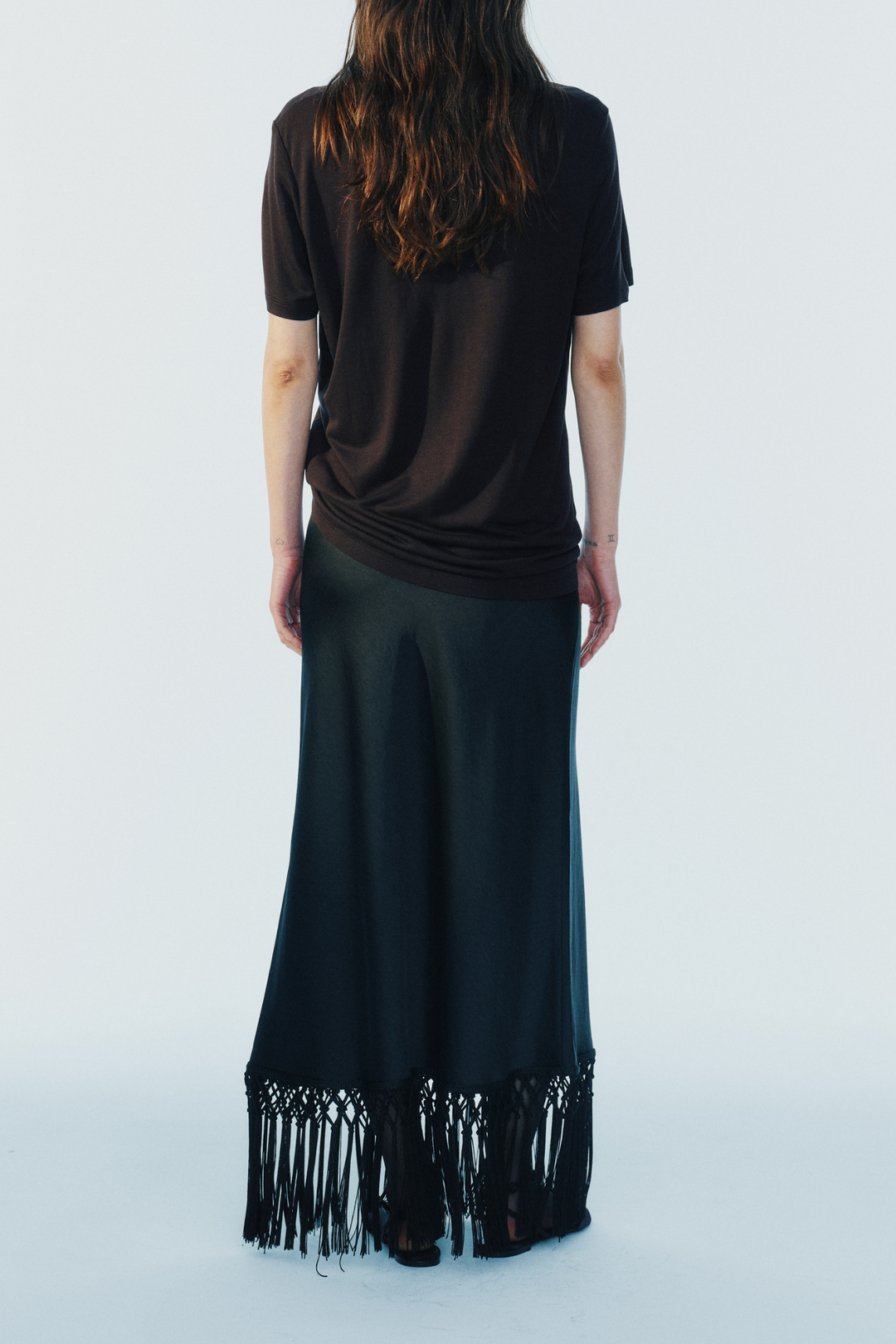 model wearing a black maxi skirt with fringe trim and a short sleeve brown tee on a white background, back view