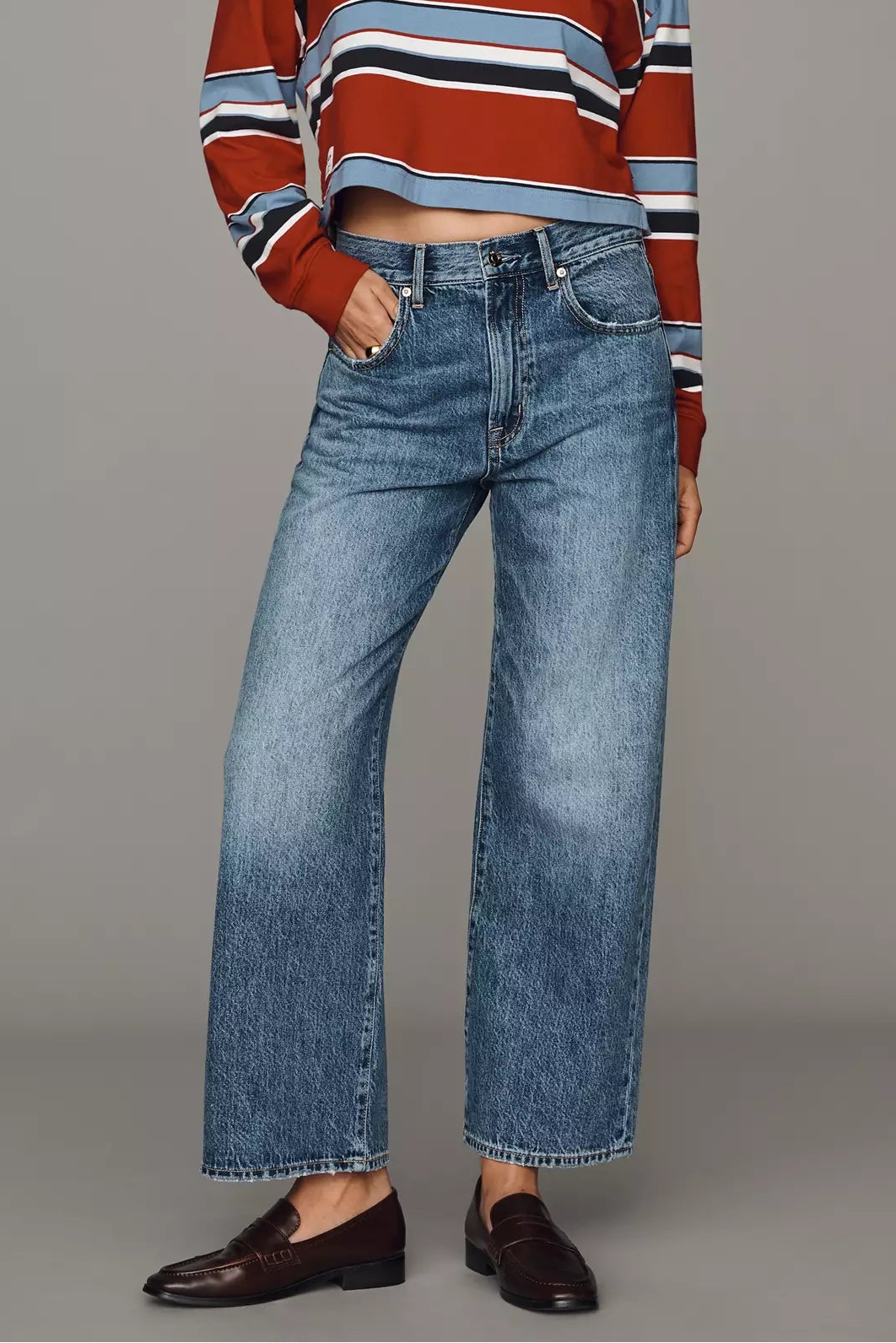 A model stands against a solid gray studio background wearing a cropped, long-sleeve striped knit polo shirt in rust red, navy, white, and light blue, paired with wide-leg blue jeans and dark brown leather loafers.