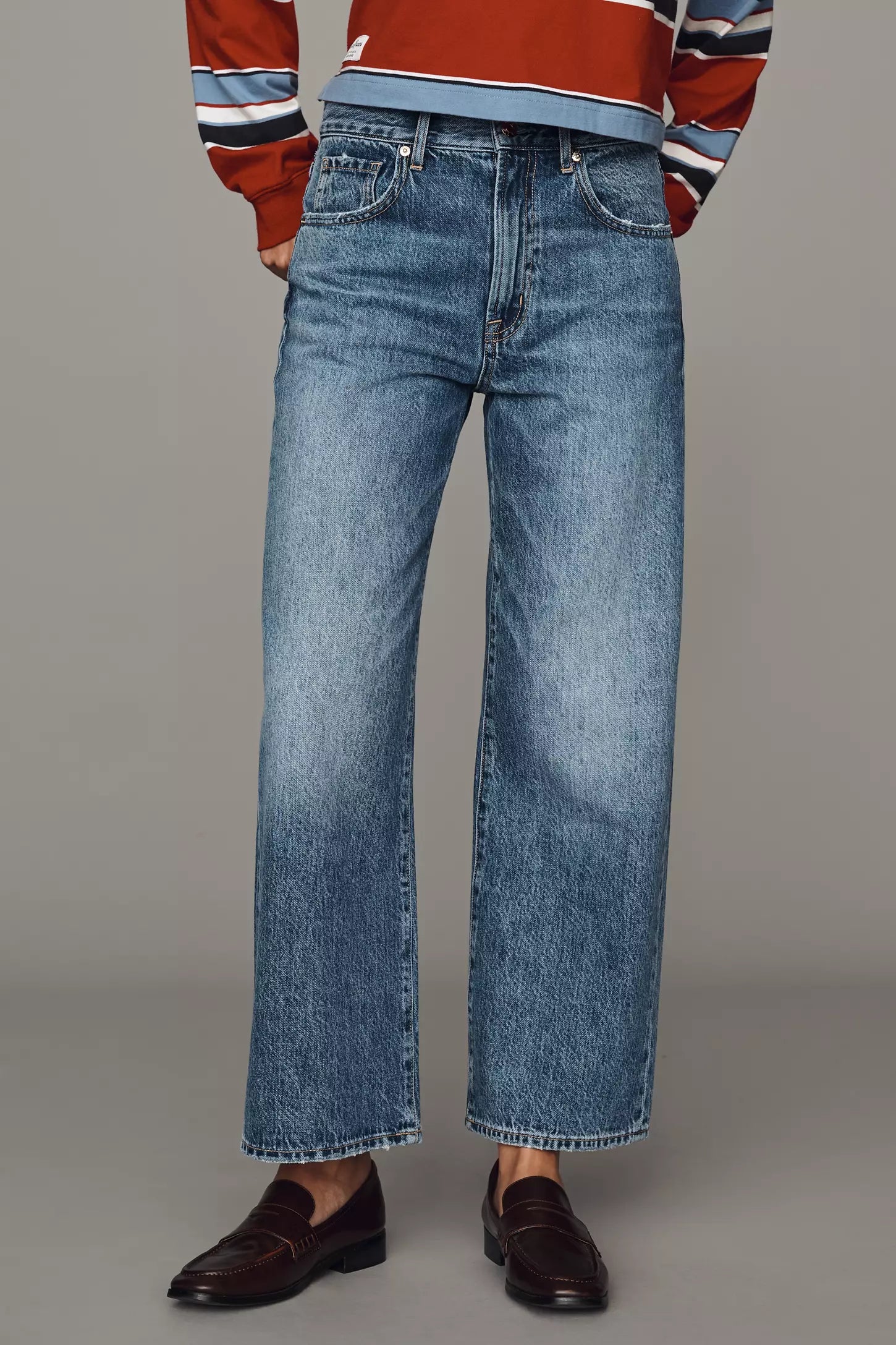 A model stands against a solid gray studio background wearing a cropped, long-sleeve striped knit polo shirt in rust red, navy, white, and light blue, paired with wide-leg blue jeans and dark brown leather loafers.