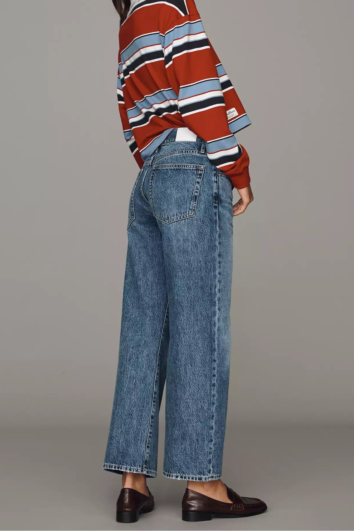 A woman modeling a rust red, navy, and white striped knit sweater paired with wide-leg blue jeans and dark brown leather loafers, photographed against a solid gray studio background.