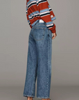 A woman modeling a rust red, navy, and white striped knit sweater paired with wide-leg blue jeans and dark brown leather loafers, photographed against a solid gray studio background.
